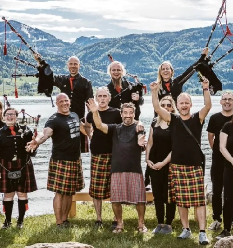 Gruppe von Dudelsackspielern in Kilts und schwarzen Uniformjacken posiert ausgelassen am Seeufer im Salzkammergut, im Hintergrund Berge und ein ruhiger See.