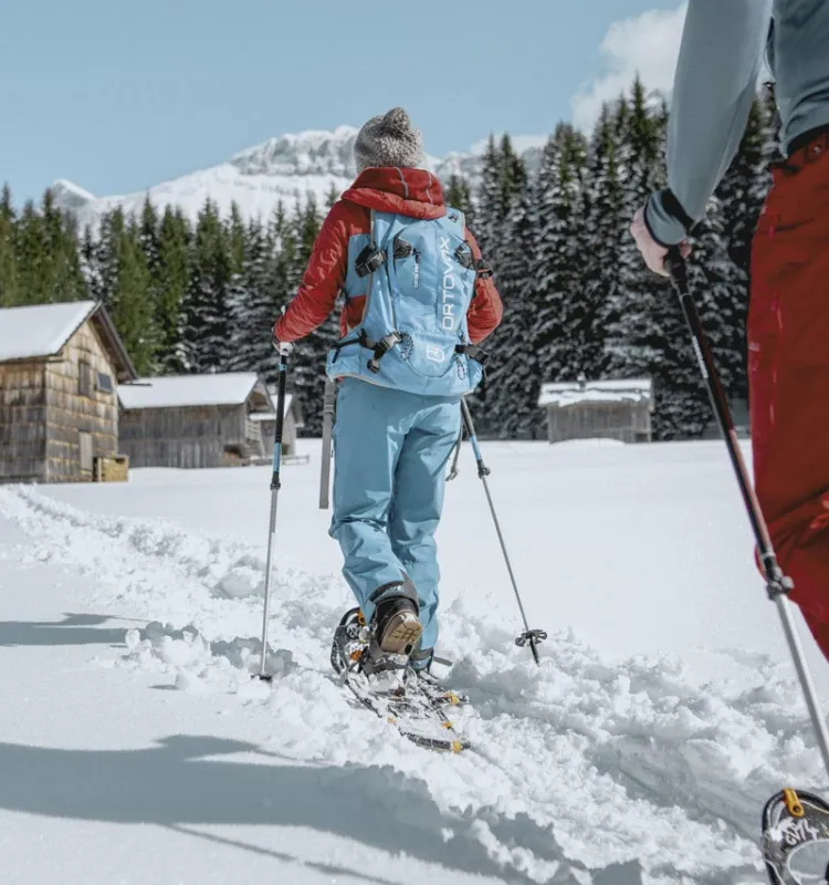 Zwei Personen beim Schneeschuhwandern durch verschneite Landschaft mit Almhütten und Bergen