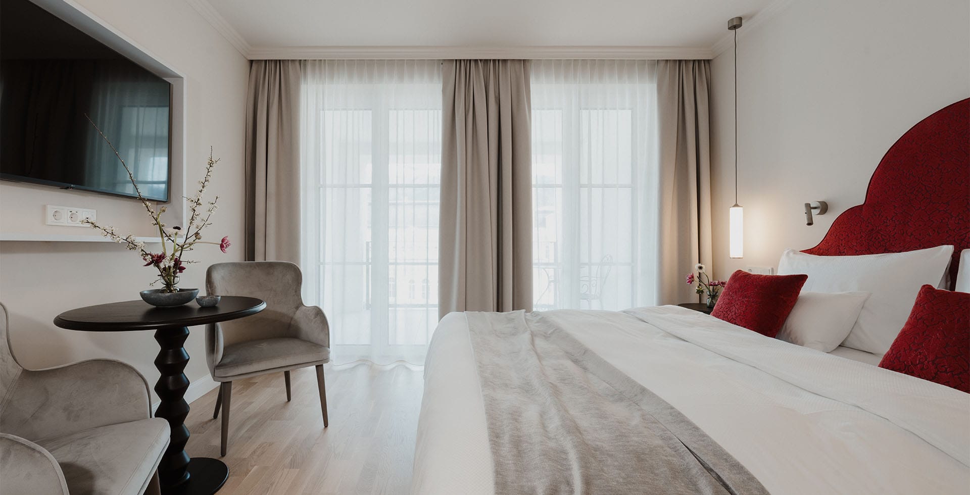 Classic Doppelzimmer mit Bett, roten Zierkissen, Sitzecke und Balkon im Grand Elisabeth Hotel in Bad Ischl.