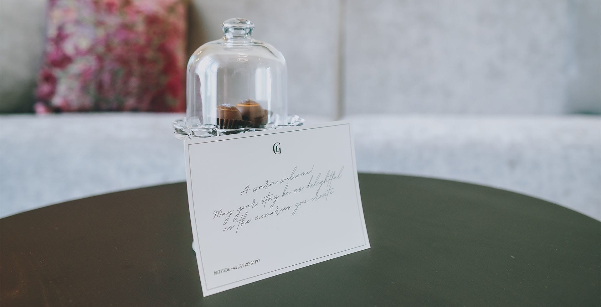 Begrüßungskarte mit feiner Handschrift und Glasglocke mit Pralinen in der Sisi Suite des Grand Elisabeth Hotel in Bad Ischl als Willkommensgruß.